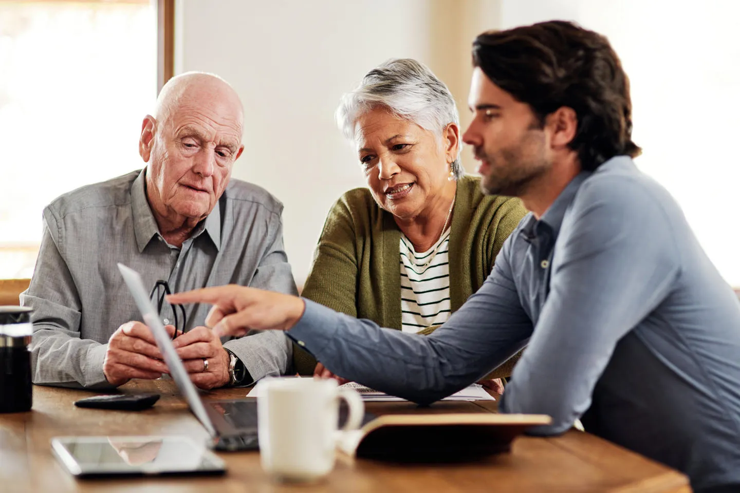 Two elderly patients sit at a table with a researcher and discuss patient involvement in research, looking at his laptop